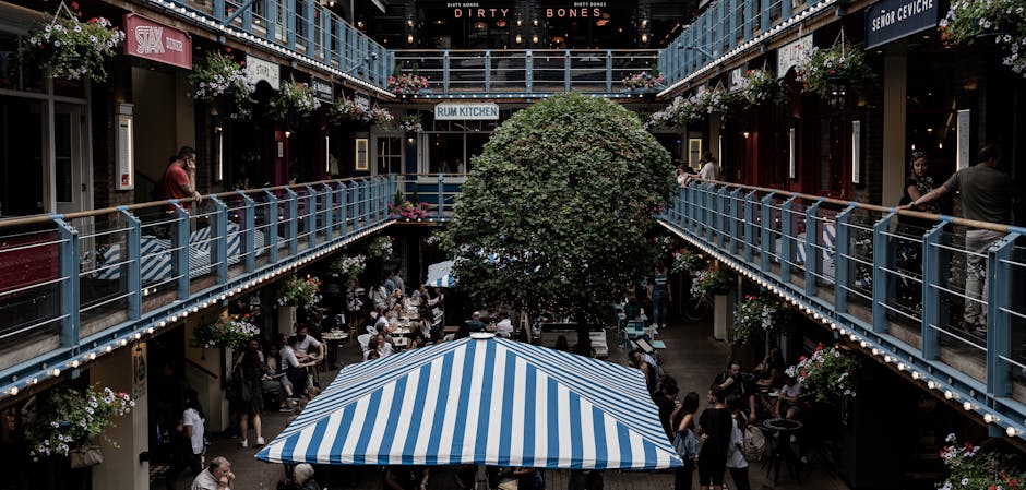 A vibrant outdoor courtyard featuring a central, lush green tree surrounded by outdoor seating under a large blue and white striped canopy. The courtyard is enclosed by two levels of shops and restaur